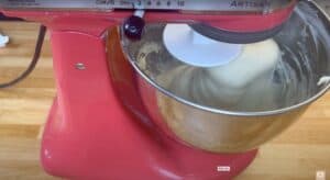 Red stand mixer kneading dough in a stainless steel bowl on a wooden countertop.